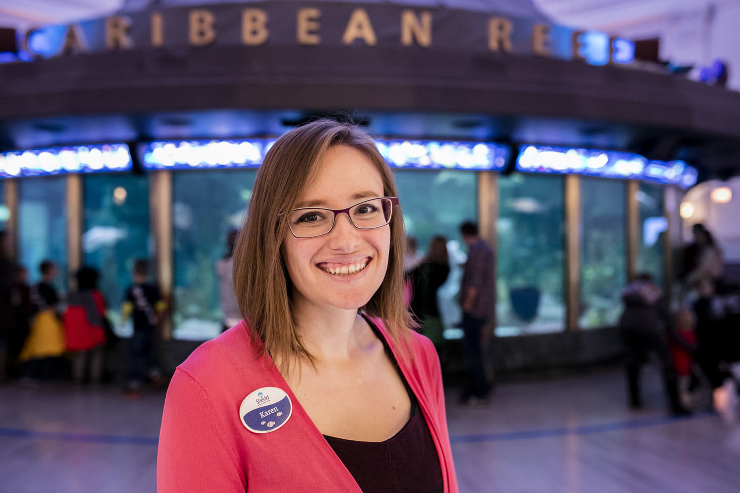 Karen is white person with shoulder-length straight brown hair, metal-frame glasses, and a bright pink cardigan stands in front of a circular 90,000-gallon aquarium with the words CARIBBEAN REEF in large lettering around the top. They are wearing a Shedd Aquarium nametag.