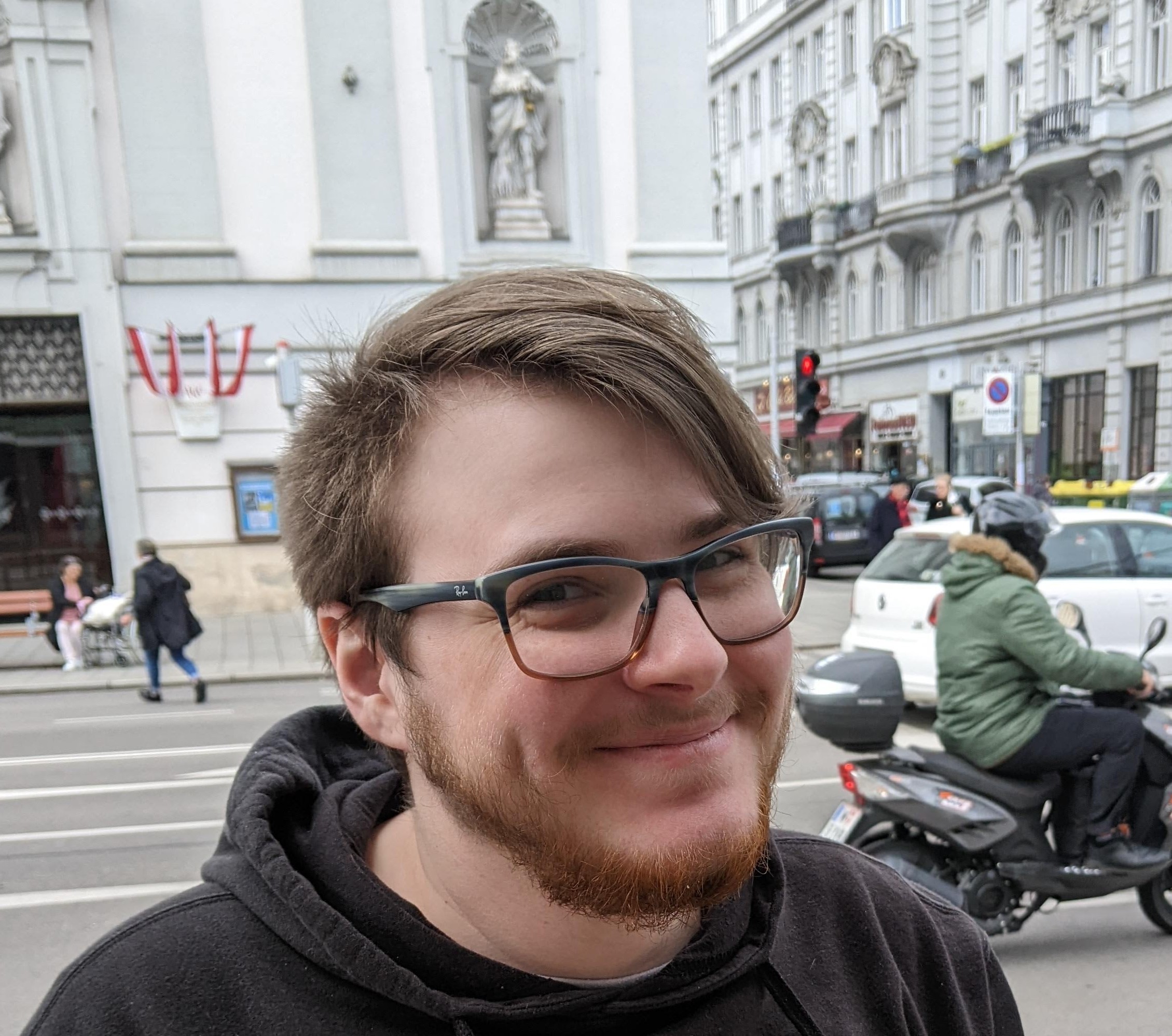 Andy is a white man in his mid-thirties with dark blonde hair, cut short on the sides and grown long in the middle and pushed to the side. He has round cheeks, deep dimples and blue eyes squinted in a smile. He has a short beard that is reddish brown. He wears glasses with blue and brown frames.