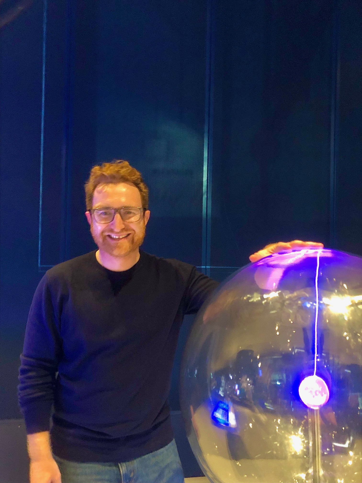 Nick is a white male with medium length red hair and a matching red beard and full rimmed eye glasses. He is wearing a blue sweater and jeans. He is at the Museum of Science and Industry standing next to a glass plasma ball with his left hand on the glass ball.