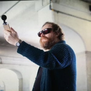 Andy extends his left hand up to the sky. He is holding a Recording device with an attached microphone. The microphone has a small, black foam ball covering it. The sun shines down on Andy’s oversized amber sunglasses, bushy red beard, and thick green sweater. Photo credit: Charlie Simokiatis. 