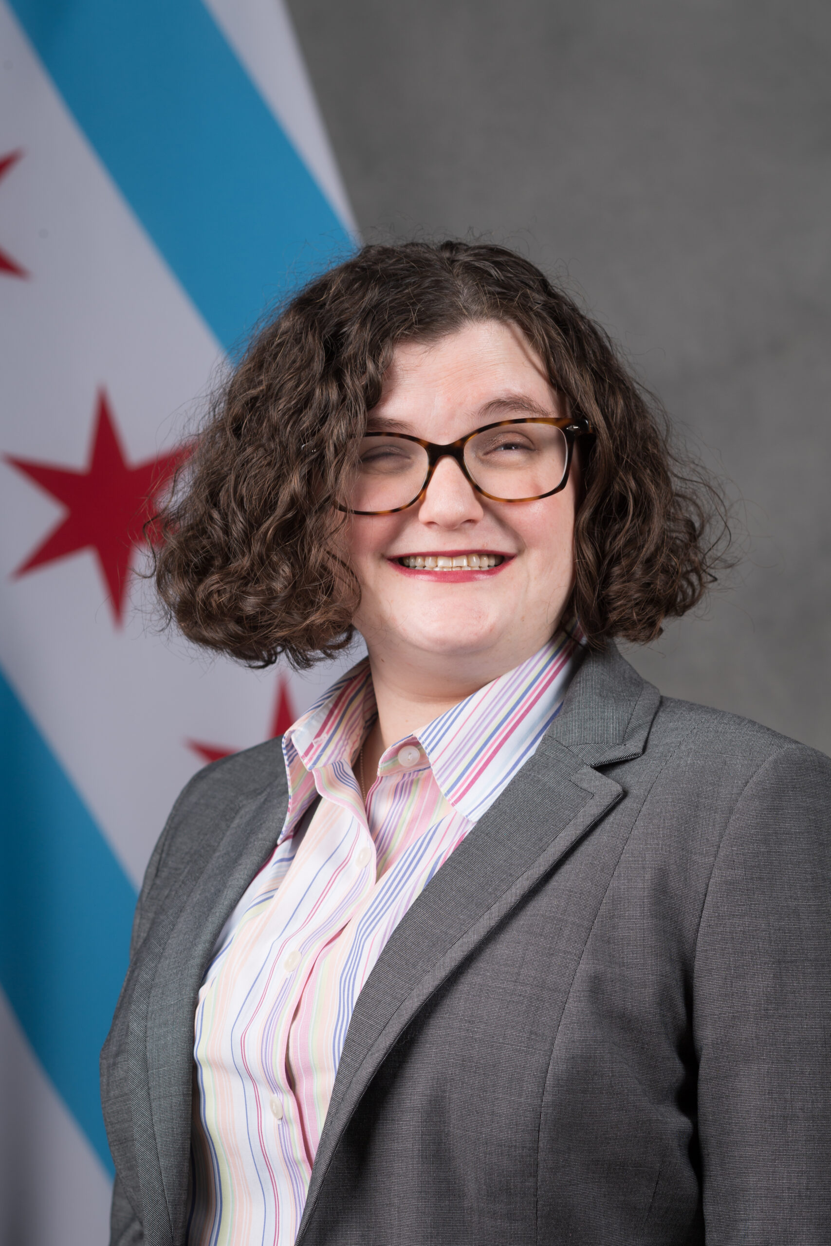 Carly is a Caucasian woman with short brown hair, wearing glasses, a collared shirt, and a gray jacket. She stands in front of a gray background with the city of Chicago flag in the background.