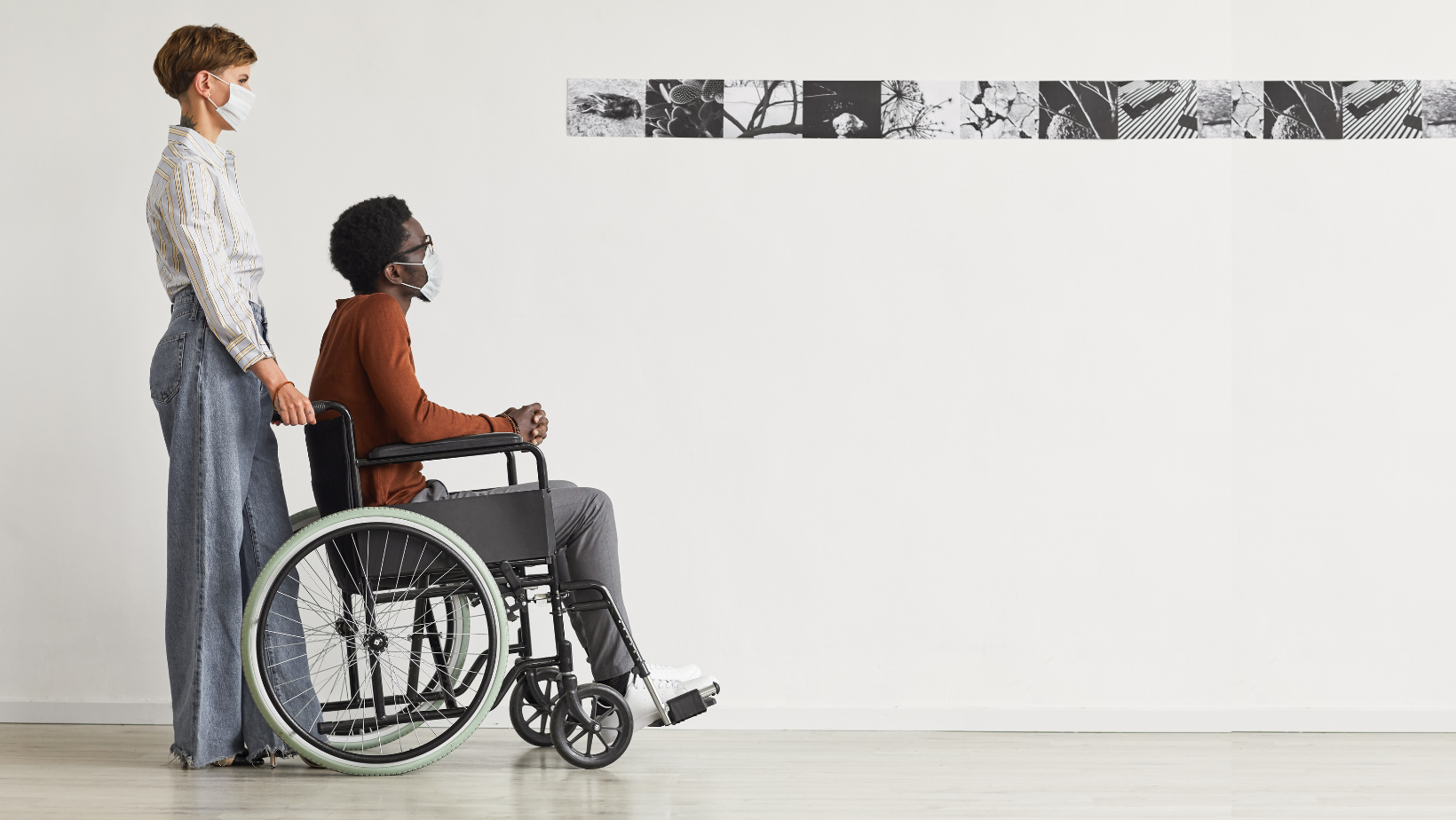 A Black man using a wheelchair looks at photos in a modern art gallery. A white woman stands behind him, hands resting on the chair's push handles. Both wear masks.