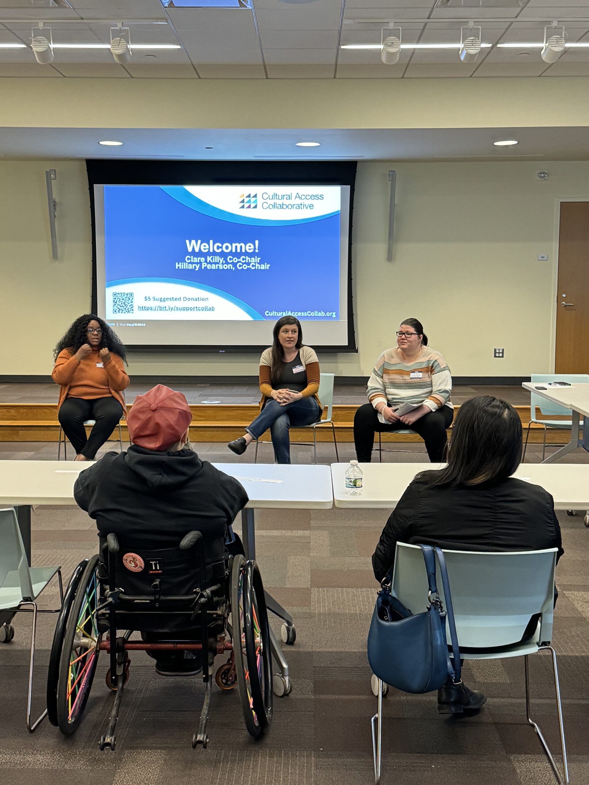 Collab co-chairs Clare Killy and Hillary Pearson, two white women wearing comfortable sweaters, sit in the center of a conference room. In the foreground, two program registrants are seated at a table with their backs facing us. The person at left is seated in a manual wheelchair with rainbow-colored spokes. The person at right has hooked a blue purse onto the back of their chair. They both listen to the presentation, as an African American ASL Interpreter signs. Behind the presenters is a projection screen with a slide that reads, “Welcome!”