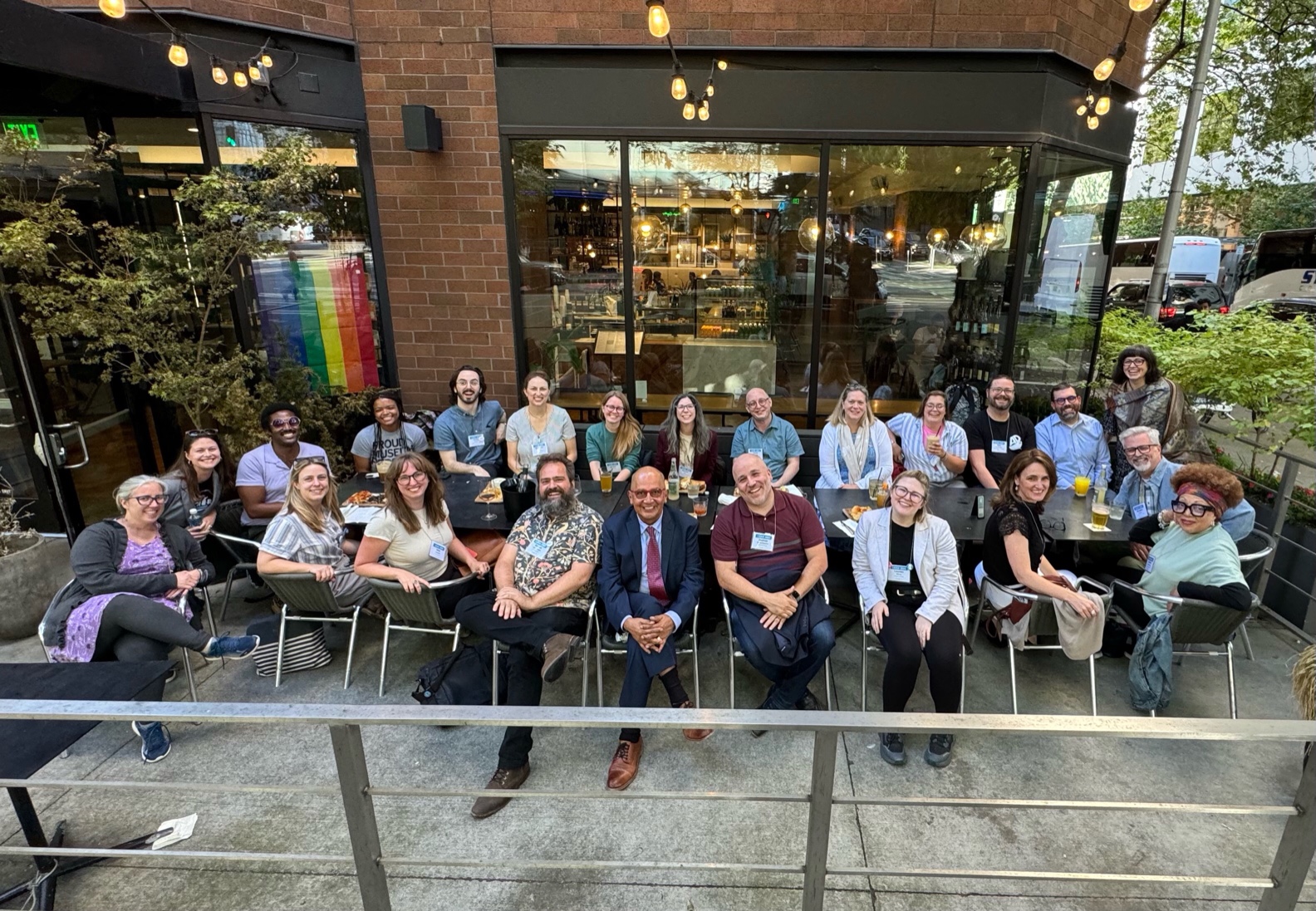 23 humans are seated around a long table on a patio outside a coffee shop. We're smiling broadly towards the camera.