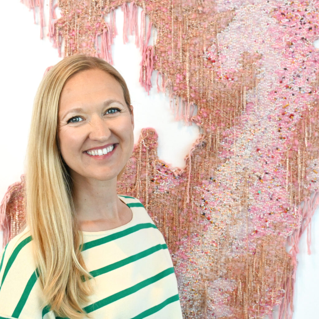  headshot of Portia Brown, a white woman with long blond hair standing in front of a woven art piece smiling