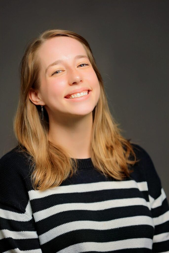Mikaela is a white woman in her mid-30s, with shoulder length brown hair and a big smile on her face. She wears a black and white striped sweater and poses against a grey background.