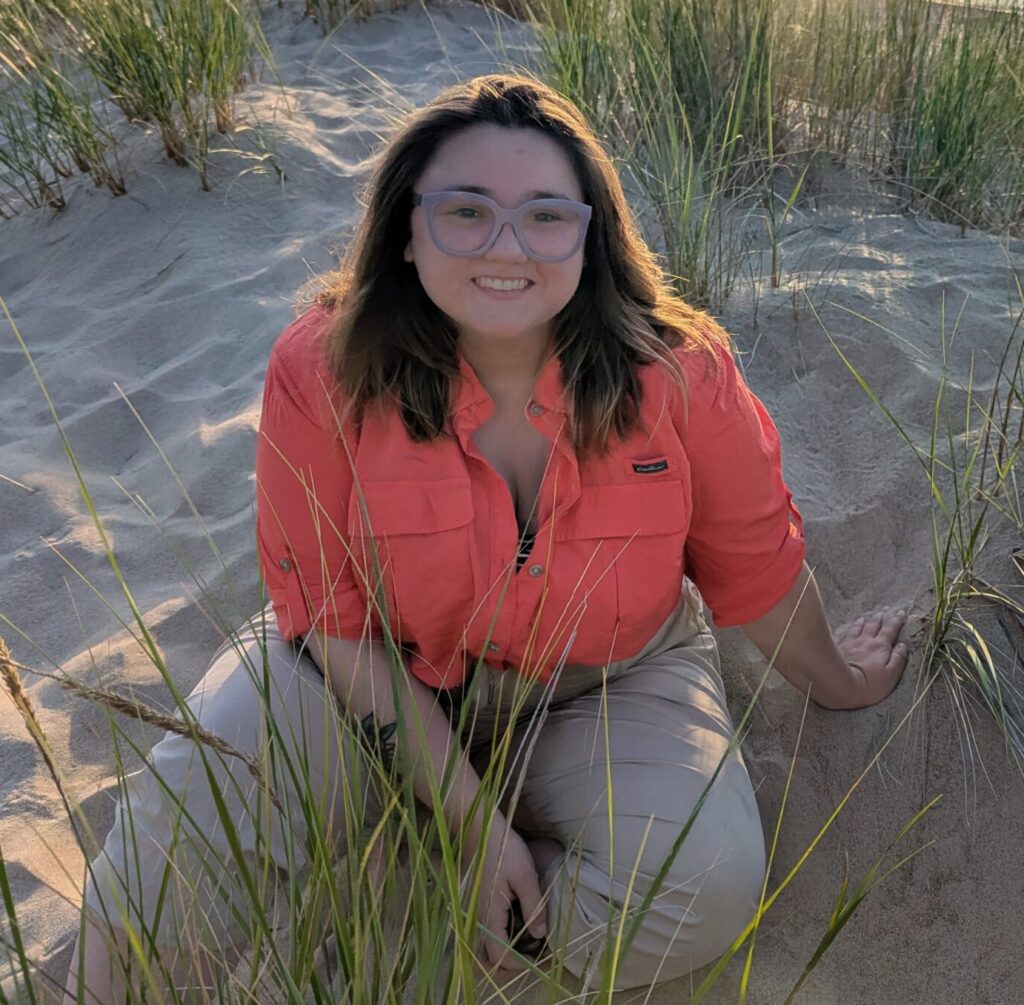 I am a white, cisgender woman in my 30s with brown eyes, shoulder-length, straight brown hair, and purple glasses. In this picture I am sitting on a grassy sand dune with Lake Michigan in the background. I am wearing an orange button-down shirt and khaki hiking pants.