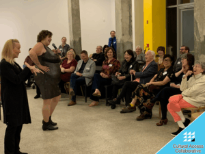 KT Zane stands in front of laughing audience in a sparkly dress with a mic. ASL Interpreter stands beside. 