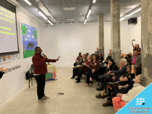 Ben Noble stands in front of a laughing audience in a red jacket holding his mic in one hand and holding his other hand out in a "stop" motion.