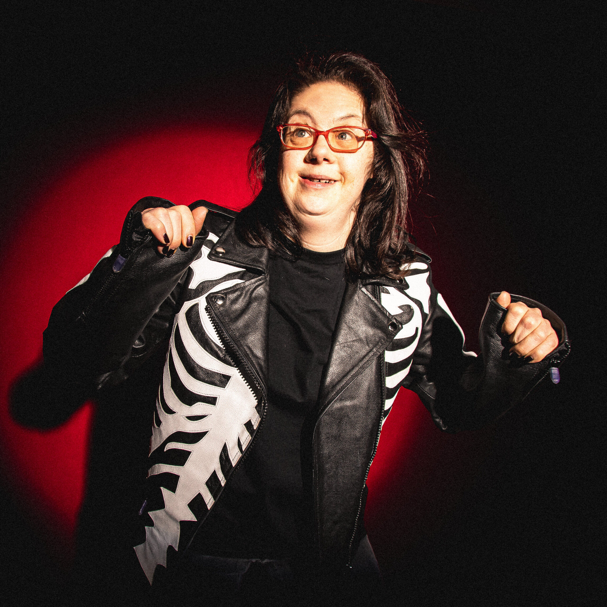 Erin stands in shadowy lighting infront of a blood-red backdrop that matches the frames of her glasses. She looks up and off camera with an expression of excited anticipation as she pumps her fists. Her dark hair moves as if she is jumping for joy. She wears a black leather jacket with silver bones emblazoned on it.