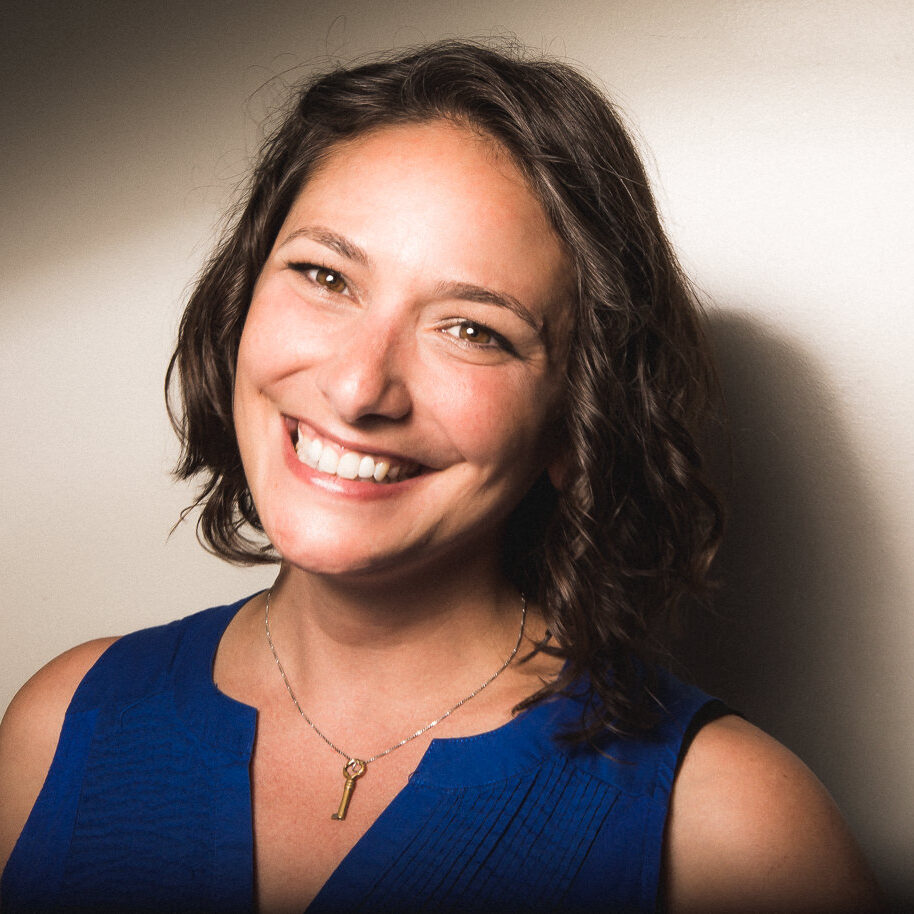 Katie, a white woman with a non-apparent disability smiles for the camera infront of an off-white backdrop in dramatic lighting. Her brown, shoulder-length hair hangs in loose curls. She wears a royal blue sleeveless top with a notched collar that reveals a necklace with a small silver key.