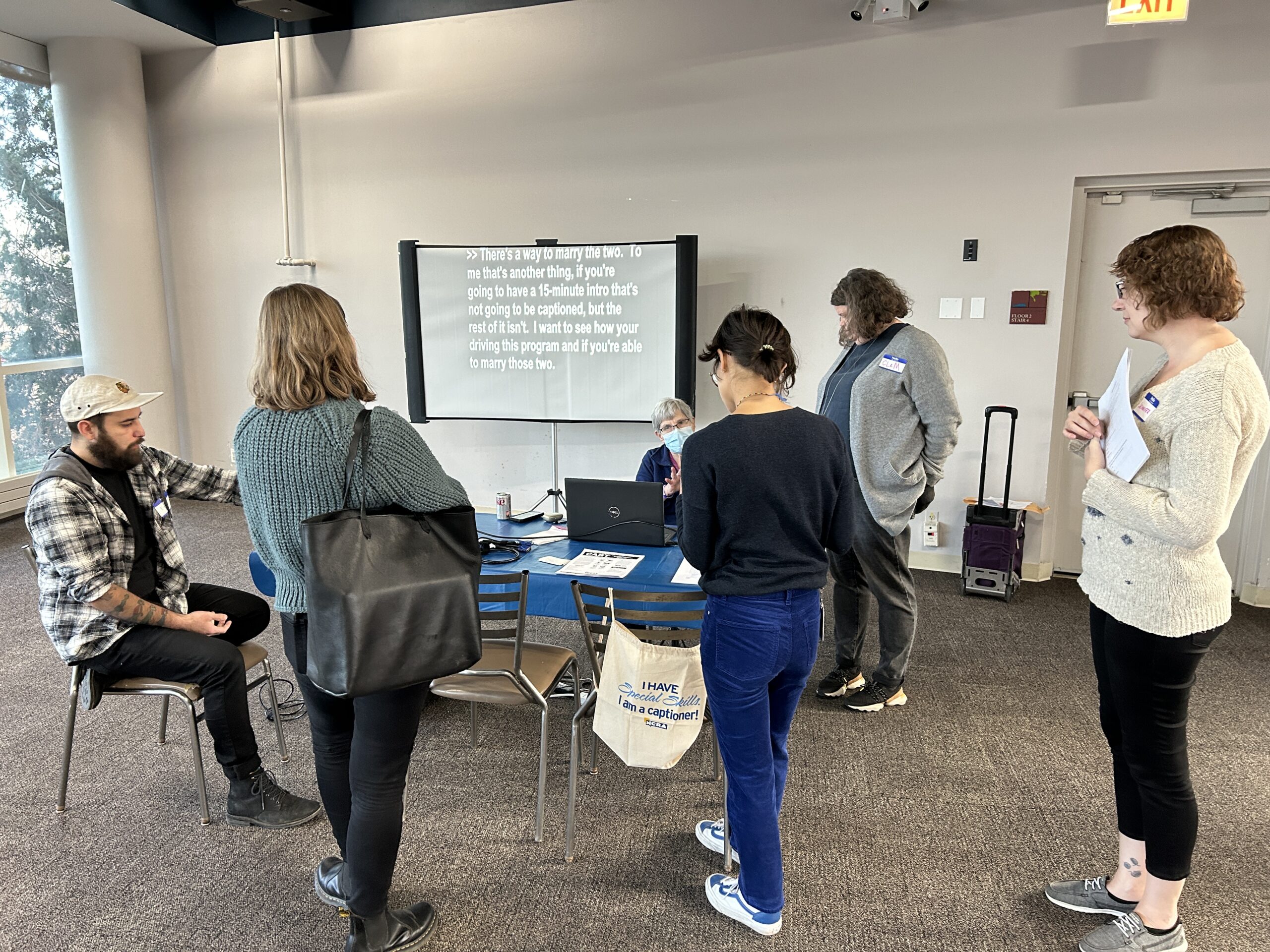 Guests stand around a table watching a captioning demonstration at the last Equipment Loan Open House