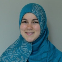 A woman wearing a blue hijab with a floral pattern smiling against a neutral gray background. 