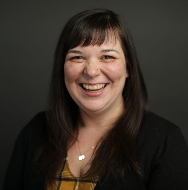 Dani Rose, a white brunette with bangs smiling big wearing a plaid dress and black cardigan against a gray background.