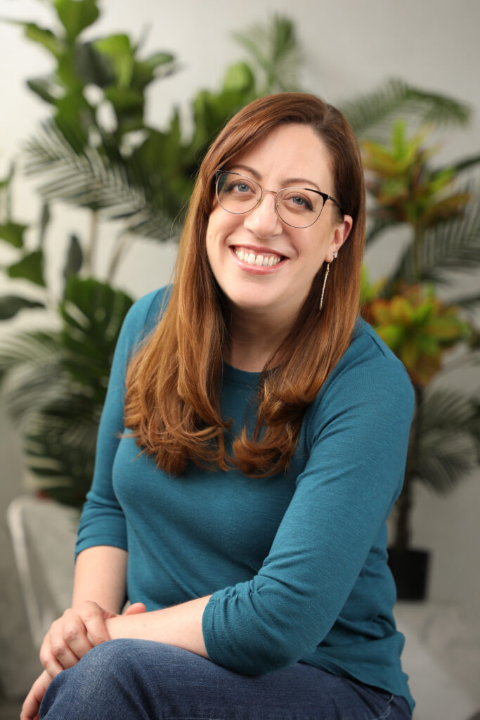 Allison Price is a white middle-aged woman with long reddish-brown hair. She is wearing glasses and a blue shirt and is seated in white room with a green-leafed potted plant behind her.  