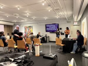 Photo of SC member Daria Smith presenting to guests seated in rows. Beside her on either side is an ASL interpreter and caption screen.