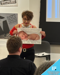 Liliana holds a baby photo up in front of the audience with a knowing look on her face.