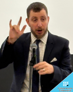 Brad stands in front of a mic in a suit, signing and speaking to the audience.