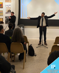 Brad stands in front of the audience posing his arms like a clock. An ASL Interpreter stands to the side.