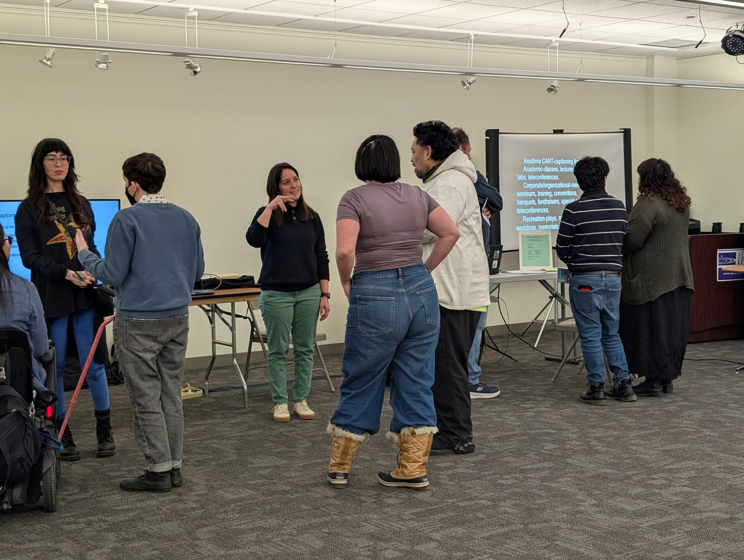 A group of people standing and talking in front of the projector and television caption kits.